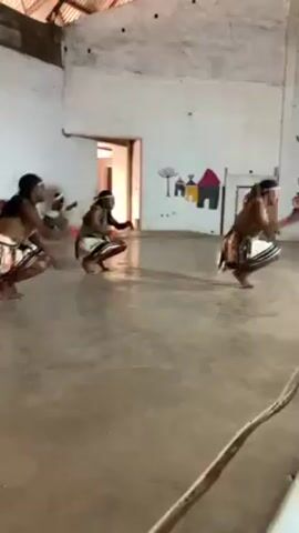 Several people are dancing in a large room. The dancers, dressed in matching skirts and headdresses, are performing a coordinated routine. They move energetically, bending low and raising their arms. The room has a concrete floor and white walls. There's a drawing of houses and a tree on one wall.  The roof appears to be made of corrugated metal. The overall impression is of a traditional performance in a simple, indoor space.