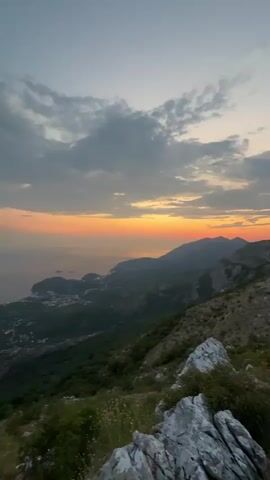 A vertical video captures a panoramic view of a mountainous coastal landscape at sunset. The sky is a gradient of pale blue at the top, transitioning to soft pink and orange hues near the horizon, with scattered clouds. The sun is setting over a calm body of water, likely the sea, which stretches to the horizon. In the middle ground, a lush green forested mountainside slopes down towards the coast. A small town or village with scattered buildings is visible nestled within the greenery and along the coastline. In the foreground, rough, greyish-white rocks are prominent, with some green foliage and dry grass growing around them. The camera pans slowly from left to right, revealing more of the expansive coastline and the distant mountains. The overall impression is one of natural beauty and tranquility during the golden hour.