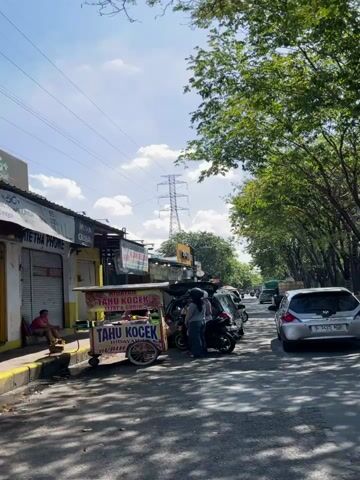 The video depicts an outdoor scene on a sunny day. In the foreground, a food cart labeled "TAHU KOCEK" is visible. Behind the cart, several people are interacting near parked vehicles, including a motorcycle and cars. One person is putting items into the trunk of a car, while others stand nearby. To the left of the food cart, a man is sitting on a low stool outside a building with various shopfronts. The background shows a tree-lined street with cars driving by and a large electrical tower in the distance. Power lines are strung across the sky. The overall impression is a bustling street scene, likely in a commercial area.