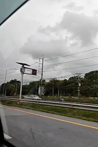 The video shows a view from a vehicle driving on a road. A traffic light with a red X is visible on the right side of the road. There are power lines running overhead and trees lining the side of the road. Several cars are driving on the road. The sky is overcast.