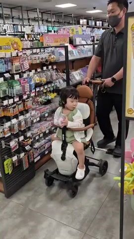 A young child sits in a stroller as a man pushes it through a store aisle. The child holds a small white object in their hands. Shelves stocked with various products line the walls of the store. The man is wearing a dark gray polo shirt and black pants. The child is wearing a light green shirt and white Crocs.