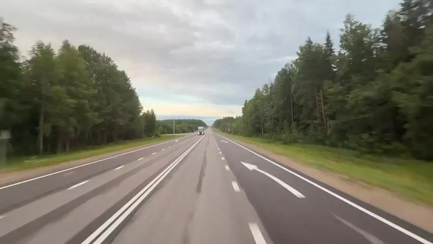 This video shows a point-of-view shot of a highway driving through a forest. The road is paved and has two lanes in each direction, separated by a median. White lines mark the lanes, and there are some faint tire marks on the road. On the left side of the road, there is a forest with tall, green trees. On the right side of the road, there is also a forest, but the trees are denser and appear taller. The sky is cloudy, with some patches of blue visible. A white truck is seen driving in the distance in the opposite lane. The sun appears to be setting or rising, casting a soft light on the scene. There are no people visible in the video, and no other vehicles are seen except for the truck. The video appears to be shot during daylight hours.