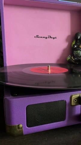 A close-up shot shows a purple "Sunny Days" record player with a black vinyl record spinning on its turntable. The record has a red label. The record player's case is open, revealing the words "Sunny Days" printed in black on a light pink interior. A small, dark-colored statue of a pregnant woman sits next to the record player, slightly out of focus. The background is dark and indistinct.