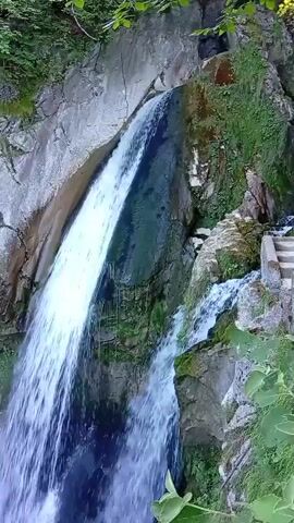 A powerful waterfall cascades down a rocky cliff face. The water is a vibrant white as it plunges into a serene, greenish pool at the bottom. Lush green vegetation clings to the sides of the cliff, adding to the natural beauty of the scene. Sunlight filters through the trees, illuminating the water and creating a shimmering effect. A set of stone stairs can be seen ascending the cliff to the right of the waterfall, suggesting a potential viewing area or pathway. The overall impression is one of untouched natural splendor and the raw power of water.
