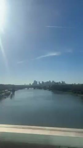 A body of water with a skyline on the horizon under a sunny blue sky. The water is calm and reflects the sunlight. There are two bridges visible in the distance.  There is a white contrail in the sky. The camera is static and the scene is captured from an elevated position, possibly from another bridge or tall building. A railing is partially visible at the bottom of the frame.