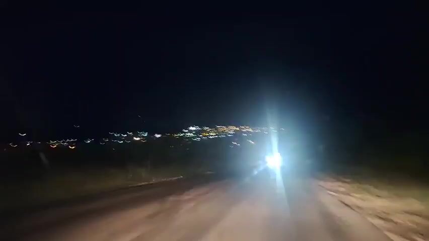 The video captures a nighttime view from the perspective of someone driving on a road towards a distant town illuminated by numerous lights. The headlights of the vehicle in front are bright and create a glare. The road surface appears to be asphalt and is wet in places, reflecting the light. The surrounding environment is dark with some vegetation visible on the sides of the road. The town lights are spread out on a gentle slope in the distance, suggesting a populated area.