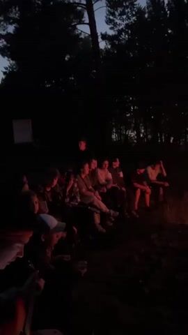 A group of people are sitting around a bonfire in a wooded area at dusk. The fire is the primary light source, casting an orange glow on the faces of the people and the surrounding trees. The sky in the background is dark with hints of pink and orange from the setting sun. The people are sitting on the ground and on logs, facing the fire. Some individuals appear to be talking and gesturing, while others are looking into the flames. The atmosphere appears to be relaxed and social.