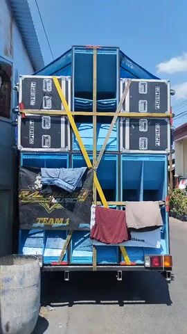 This video shows the rear of a blue truck loaded with large speaker cabinets. The cabinets are stacked high and secured with yellow straps and wooden beams. A black banner with white lettering that reads "RASYID MUSIC TEAM 1" is attached to the lower section of the speaker stack. Several pieces of clothing, including a maroon shirt and a grey garment, are draped over the speaker cabinets. Two people on scooters are visible in the background, riding on a paved road alongside the truck. The environment appears to be a sunny day in a suburban or rural area, with houses and vegetation visible along the roadside.