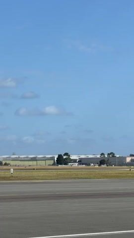 A white airplane with red and white checkered tail livery and "VOLOTEA" written on its fuselage taxis from right to left across a paved airport surface. The aircraft is visible in the midground. In the foreground, a grey asphalt runway or taxiway with yellow and white markings is empty. Behind the airplane, a strip of green grass leads to distant low-rise buildings, possibly hangars, and scattered trees. The sky is mostly clear blue with a few white clouds concentrated on the left side of the frame. The scene is outdoors on a bright day.