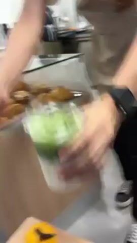 The video shows a person placing a plastic cup containing a layered drink on a small table next to a display case of croissants. The drink appears to have a white layer at the bottom and a green layer on top, possibly a matcha latte. The person places a straw next to the cup. The video then shows two people behind the counter of the bakery. Both are wearing aprons and t-shirts. One is working at a register. The other is walking toward the back.