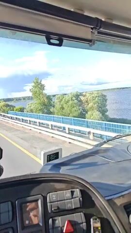 The video is shot from the perspective of someone inside the cab of a truck, looking out through the windshield. The truck is driving on a road that appears to be a bridge or causeway over a body of water. On the right side of the road, there is a blue and white guardrail, and beyond that, lush green trees and bushes line the water's edge. The sky is bright blue with fluffy white clouds, and the sun is shining. In the distance, more trees and what appears to be a shoreline can be seen. The truck's dashboard and steering wheel are partially visible in the foreground. A small, square electronic device with a digital display is mounted on the dashboard. There are also some buttons and controls visible on the dashboard. The road has a yellow line marking the lane. The truck is moving forward, and the scenery is passing by.
