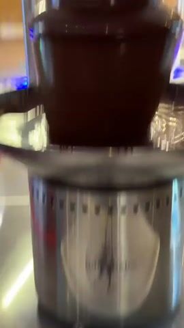 The video shows a close-up view of a chocolate fountain. The fountain has multiple tiers, and melted chocolate is flowing down the sides. The chocolate appears to be dark brown and glossy. The fountain is on a silver base with ventilation holes. In the background, there are glimpses of a brightly lit environment, possibly a restaurant or buffet, with out-of-focus objects and reflections. The lighting is warm, creating a festive atmosphere. The camera movement is subtle, focusing on the cascading chocolate.
