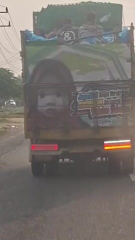 A truck with a green tarp covering its load is driving on a paved road. The back of the truck is decorated with a large image of a child's face and colorful graffiti. Three people are visible in the back of the truck, sitting on top of the load. One person is wearing a black shirt with the number "8" on it. The truck is in motion, with trees and foliage visible on the side of the road. The sky is overcast.