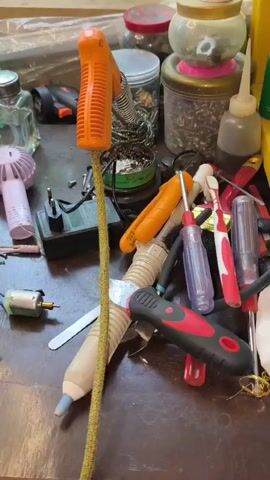 A bird's-eye view of a cluttered workbench shows various electronic components and tools. In the foreground, a disassembled blue plastic device reveals its internal circuitry, including a small green circuit board with a lit LED and a connected motor. A USB cable links the circuit board to the device's housing. Nearby are loose screws, a metal blade attachment, and a white plastic piece. To the left, a multimeter with red and black leads is connected to an unknown device. A small pink fan sits behind the multimeter. In the center of the frame, a long, yellow, braided cable with an orange handle extends vertically. Various other tools and components are scattered around the table, including pliers, screwdrivers with orange and red handles, and jars containing screws and other hardware. A bottle of clear liquid and a pink toothbrush are also visible. The lighting suggests an indoor environment, possibly a workshop or repair area.