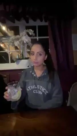 A woman with long dark hair in a ponytail sits at a wooden table in a dimly lit room. She is wearing a gray sweatshirt with "Athletics Club" printed on it. She holds a glass filled with ice and liquid, possibly a cocktail, and drinks from it using a straw.  Behind her is a window with white trim. A vase of white flowers sits on the windowsill.  A dark burgundy curtain hangs to the right of the window. The lighting is warm and seems to be from indoor fixtures.