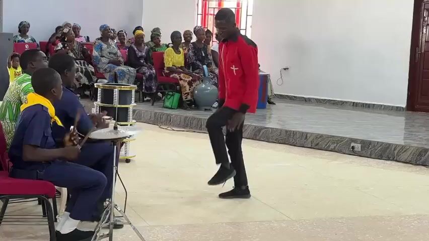 A young man in a red and black shirt and black pants dances in a brightly lit room with a light-colored floor. He has short hair and moves his feet and legs in a rhythmic way, lifting his knees and tapping his feet. To his left, three young men are seated, with two of them playing drums. The drummer on the right is playing a small golden drum set. In the background, an audience of around twenty people, mostly women, are seated on red chairs, watching the performance. Some are smiling and some are raising their hands. The room has light-colored walls, a window on the right, and a raised stage area behind the dancer.