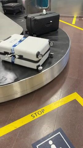 The video shows a baggage claim area at an airport. Luggage is moving on a conveyor belt. A person's hand reaches out and grabs a black suitcase. The suitcase has red stickers on it. Yellow lines on the floor indicate a "STOP" zone.