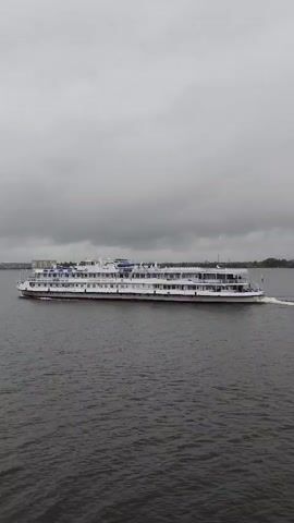 A drone shot captures a long, white multi-decked riverboat sailing from left to right across a wide expanse of dark, choppy water. The sky above is overcast and gray. The boat appears to be moving at a moderate speed, leaving a white wake behind it. The shoreline is visible in the distance on the left, with what appear to be buildings and trees. The boat has numerous windows along its sides and a series of decks stacked vertically. The overall scene suggests a cloudy day on a river or large lake.