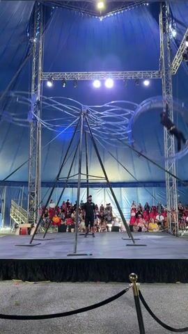A circus performer is suspended within a large metal wheel-like structure. The performer, dressed in dark clothing, is seen in mid-air performing acrobatic maneuvers. The wheel is attached to a central pole structure with multiple radial supports, creating a tripod-like base. The entire apparatus is situated on a raised stage. In the background, an audience is seated, observing the performance. The circus tent has a blue canvas ceiling, illuminated by spotlights.