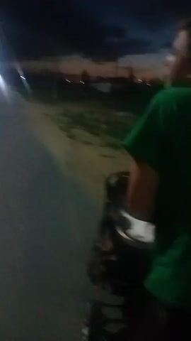 A handheld video records a nighttime scene with a road in the foreground. To the left of the road, a light pole and a silver car are visible. Further down the road, a person wearing a green shirt is riding a motorcycle. The sky in the background shows the last vestiges of twilight with a few distant lights. The camera is in motion, suggesting the video is being recorded while walking or riding. The lighting is dim, with the primary illumination coming from the light pole and the motorcycle's headlight.