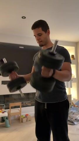 A man is performing bicep curls with dumbbells in a living room. He is wearing a dark grey t-shirt and black sweatpants. The room appears to be a home, with a large television on the wall, a fireplace, and children's toys scattered on the floor. There are also shelves with decorative items. The lighting suggests it is daytime. The man's facial expression is one of concentration.