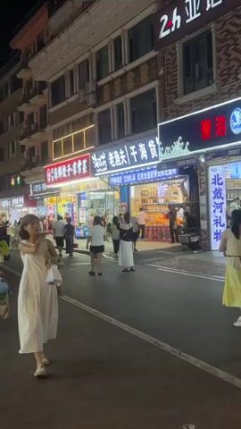 This is a nighttime, eye-level, medium shot of a street scene. A woman in a white dress walks towards the camera on the right side of the street. She is carrying a white bag in her left hand and a brown bag over her shoulder. She has short, dark hair and is looking to her right. Behind her, a man and a woman walk away from the camera. The man is wearing a white t-shirt with black lettering and black shorts with white designs. The woman is wearing a pink tank top and denim shorts. They are both holding blue objects, possibly helmets, and the man is also carrying a brown bag. The street is lined with buildings with shops on the ground floor. The shops have bright signs with Chinese characters. There are other people walking on the street and on the sidewalk. The street is paved with asphalt, and the sidewalk is made of interlocking bricks. The lighting is from the shop signs and streetlights.