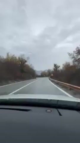 The video shows a car driving on a wet, winding road, on a cloudy day. The road is surrounded by trees with bare branches and a few trees with yellow leaves. The road has a white line dividing the lanes, and a red and white road sign is visible on the right side of the road. The car appears to be heading towards a mountain in the distance.