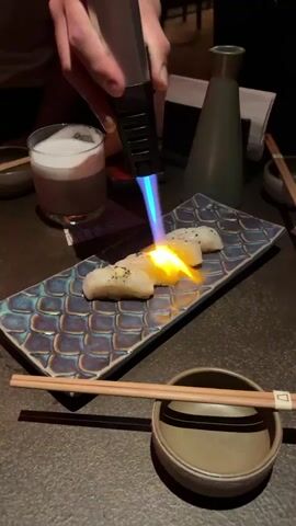 A person's hand is visible in the upper left corner of the frame, holding a culinary torch with a bright yellow and blue flame. The torch is being used to sear three pieces of what appears to be sushi or some other food item, arranged on a decorative, dark blue and grey plate with a textured pattern. The food pieces are lightly seasoned with black pepper. The plate is situated on a dark, textured surface, likely a table. In the background, to the left, is a glass with a foamy, amber-colored drink. To the right, a dark green, tapered bottle and a white napkin are visible. A pair of chopsticks rests horizontally above a small, dark bowl in the foreground. The lighting is dim, creating a moody ambiance.