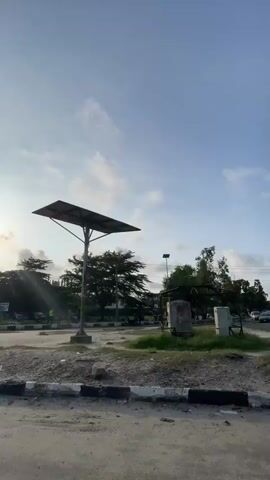 The video begins with a view of a street scene with a prominent solar-powered light pole in the foreground. The sky is clear with some clouds. A person walks into the frame from the right, wearing a blue jacket and dark pants. Another person, wearing a red shirt and black pants, walks from left to right carrying a load on their head. A blue and white van drives across the scene, followed by a red car. The camera pans to the left, showing more vehicles and buildings in the background. The sun is visible in the sky, casting long shadows.