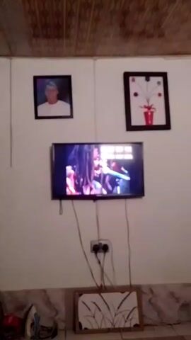 The camera shows an indoor wall with a mounted television playing a video of a person singing on a stage. Above the television are two framed pictures, one a portrait of a man and the other a drawing of a flower arrangement. Beneath the television, a power outlet is visible with cords plugged into it, and below that is a framed picture of wheat. An iron and other objects are on the floor. The ceiling has patterned tiles.