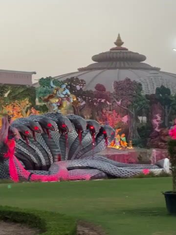 The video shows a statue of a multi-headed snake with red eyes. The statue is surrounded by other statues of people and trees. The statue of the snake is lying on the ground and there are red lights around it. In the background, there is a building with a dome. The sky is a pale pink color. The camera pans slowly.