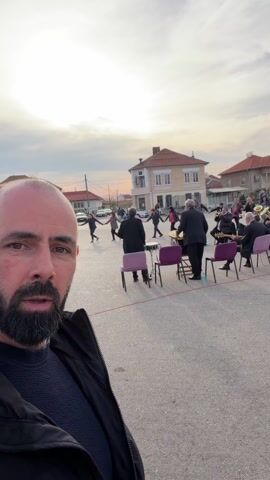 The video shows a man with a beard filming himself with a phone or camera, seemingly a selfie, in an outdoor square. Behind him, people are dancing in a circle, holding hands. A band is playing instruments in the center of the circle of dancers. They are sitting on chairs. There are buildings surrounding the square, with light-colored facades and red-tiled roofs. There are also some cars parked nearby. The sky is overcast. Another man walks past the videographer, partially obstructing the view.
