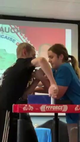 Two people are arm wrestling on a specialized table with red trim. The man on the left wears a black shirt, while the woman on the right wears a blue shirt and has her hair in a ponytail. The table has a blue block in the center and the logo "FFFORCE" printed on it. Behind them is a projector screen displaying an image with French text and a photo of a woman. There is a skull decoration on a surface below the arm wrestling table. As the wrestling begins, the woman strains visibly, and someone to the side is cheering. The scene takes place in a room with fluorescent lighting.