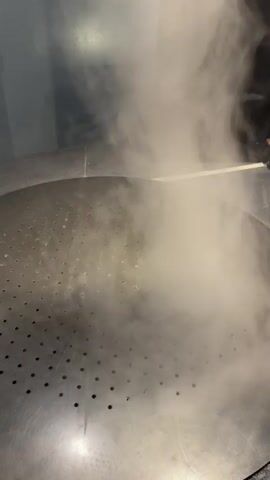A vortex of white smoke is being created above a large, circular, perforated metal surface. The smoke is concentrated in a tall, swirling column that tapers towards the bottom. A thin white object, possibly a rod or stick, is being used to interact with the smoke at the top. The background is dark, with some dimly lit walls and a black vertical object to the right. The lighting is low, creating a dramatic effect.