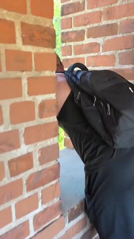The video opens with a close-up of a man wearing a black backpack and sunglasses, his back to the camera. He is partially obscured by a brick wall. The scene then transitions to a wider shot of an urban landscape with a clear blue sky. In the distance, several buildings are visible, including a pink and yellow building with many windows, a white church with a dark dome, and a taller red-and-white building. A green grassy slope with trees lines a road where cars are parked. Further down, a paved path runs parallel to the slope, with a few people walking. The overall atmosphere suggests a pleasant, sunny day in an urban park or historical district. The light indicates it is daytime, likely late morning or early afternoon. The presence of a church and older architecture hints at a location with historical significance. The people appear to be leisurely strolling, suggesting a relaxed environment. No text is visible in the video.