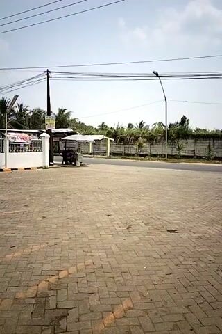 The video opens with a view of a paved area with brick-like paving stones, a street, and a low wall in the background. There is a white building with a fence on the left side, and a vendor cart under a canopy. Utility poles and wires are visible against a clear sky. Two people on a scooter pass by the vendor cart from right to left. Another scooter passes by in the distance, moving from left to right. The camera pans to the right, revealing more of the paved area, the street, and the low wall, with vegetation and trees behind it. Two more scooters are parked in front of the white building on the left. The camera continues to pan right, showing a motorcycle parked in the paved area. The lighting suggests it is daytime, with the sun casting shadows.