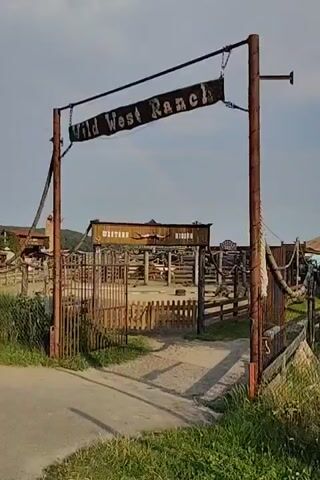 The video shows an outdoor entrance to a place called "Wild West Ranch". A rustic wooden archway with a sign that reads "Wild West Ranch" hangs over a dirt path leading into the ranch. Beyond the archway, a wooden fence encloses an area with various structures, including what appears to be a corral or arena, and a building with a sign that says "Western". There are also some trees and vegetation visible in the background. The sky is partly cloudy.