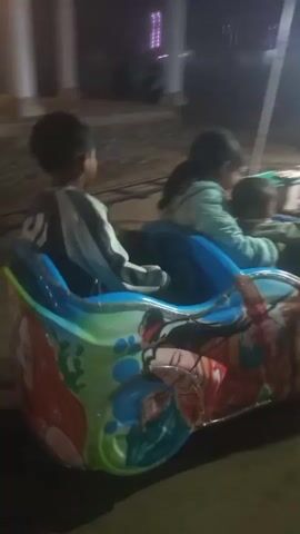 The video shows an amusement park ride with several colorful cars, each with a different cartoon character painted on the side. The cars appear to be moving in a circular motion around a central structure covered by a blue tent. Several children and adults are visible riding in the cars, some of them looking at the camera. The ground is dirt, and there are lights strung up around the tent, suggesting it is nighttime. The overall impression is of a fun, family-friendly amusement park.
