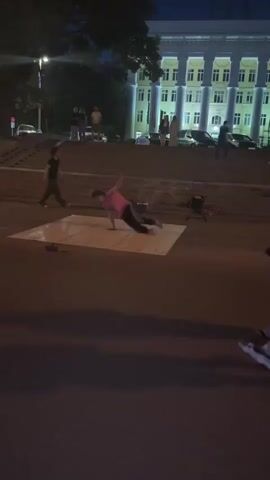 A group of people are gathered on a sidewalk at night. In the foreground, a person in a pink shirt and black pants is breakdancing on a white mat. Two people walk past the dancer on the right. In the background, a set of stairs leads up to a large, well-lit building. Several other people are standing on the stairs and in front of the building. Cars are parked in front of the building. The overall lighting suggests it is dusk or night.