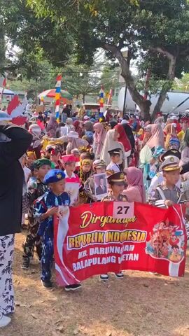 The video shows a parade or procession of children dressed in various uniforms and costumes. They are walking on a dirt path outdoors, with trees and a building visible in the background. Many adults are present, watching the children and participating in the event. Some of the children are carrying a red banner with Indonesian text and images. The children are of different ages, and some are wearing hats and carrying flags. The overall atmosphere appears festive and celebratory. The sun is shining, and the sky is visible. There are also decorations and tents in the background.