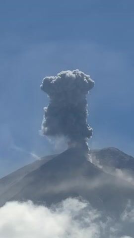 A time-lapse video shows a volcano erupting against a clear blue sky. A large plume of grey ash billows upwards from the summit of the volcano, forming a mushroom-like shape. Wisps of clouds drift around the base of the volcano.