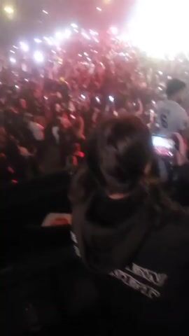 The video shows a crowded concert venue filled with people holding up their phones with the flashlights on. The stage is lit with red and white spotlights, and the performer can be seen silhouetted against the lights. The crowd is dense and appears to be enjoying the show, swaying to the music and recording the performance.