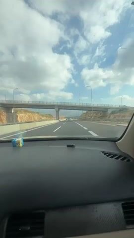 The video is shot from the interior of a car driving on a highway. The view is out of the windshield and shows a multi-lane highway with an overpass ahead. The sky is partly cloudy with bright sunshine. The dashboard of the car is visible in the foreground, with a pack of cigarettes and a black object on it. The car is moving at a moderate speed. The surrounding landscape is hilly and arid with some vegetation.