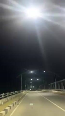 The video shows a view from a vehicle moving along an empty bridge at night. The road is illuminated by streetlights. The sides of the bridge are visible, showing metal structures. The night sky is dark, with the lights creating a starburst effect. The vehicle seems to be moving steadily along the bridge.