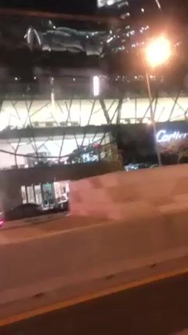The video shows a nighttime view from a moving vehicle, likely on a highway or elevated road, driving past a modern commercial building. The building has distinctive architectural details, including a bridge-like structure connecting different sections. Signage for brands such as Cartier and "L.T.K. KAI TAK" are visible on the building. Streetlights illuminate the scene, and the camera captures the concrete barriers of the roadway as it passes by. The environment is urban, with a focus on the commercial architecture and transportation infrastructure.
