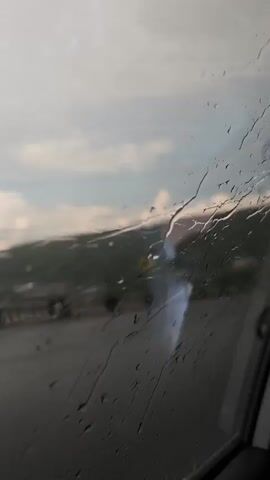 The video is filmed from inside a vehicle, looking out the passenger side window. Raindrops are visible on the window, distorting the view of the outside environment. The outside scene is blurry but appears to be a landscape with hills or mountains in the background, covered in greenery. In the middle ground, there is a road or highway, and on the left side, a yellow sign with a red circle and black symbol is visible. A tall, dark structure, possibly a utility pole or pylon, is also visible in the distance. The sky is cloudy with patches of lighter sky suggesting daylight. The overall impression is of a cloudy, possibly rainy day, viewed through a wet window while traveling.