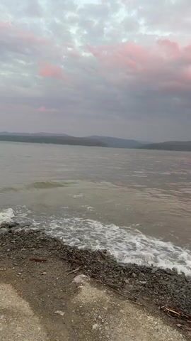 A view of a body of water with a rocky shore and a cloudy sky during sunset. The water is choppy, with small waves crashing onto the shore. The sky is filled with gray clouds, with some hints of pink and purple from the setting sun. Distant hills can be seen on the horizon. There are no people or man-made objects visible in the video.
