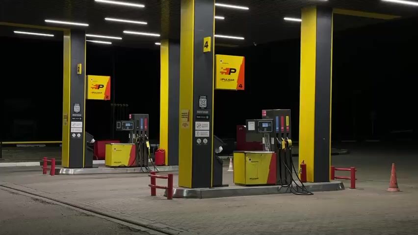 The video shows a gas station at night. Two gas pumps are visible, illuminated by overhead lights and the screens on the pumps. Yellow and black signage is present on the pillars supporting the canopy. In the foreground, a person with short, dark hair is seen walking from left to right, with their back to the camera. The ground is paved with interlocking bricks. The overall atmosphere is dark and still.
