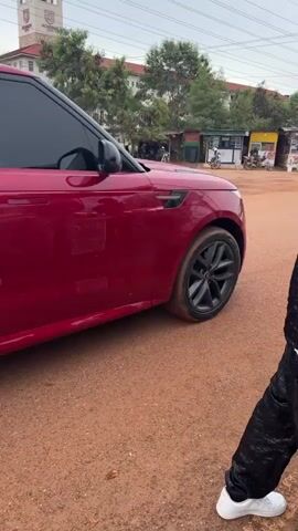 A person wearing white sneakers and a black and white ensemble walks past a dark red Range Rover Velar parked on a dirt lot. The Range Rover has tinted windows and black rims and is facing away from the viewer. In the background, buildings, trees, other cars, and a small food stand are visible. The sky is overcast.