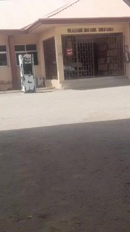 The video shows a building with the sign "HHH ALI FARUK UMAR FARUK EMIR OF DAURA".  The building is single-story, with a light-colored exterior and a reddish roof. It has barred windows and a gate in the front. There is a beverage cooler or vending machine to the left of the entrance, and a motorcycle is parked to the right. The ground in front of the building is paved. The video is somewhat shaky and appears to have been taken from a vehicle. The light suggests it is daytime.  The environment is generally quiet and there are no visible people.